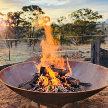 Load image into Gallery viewer, 1800mm cast iron Fire pit with flames burning in an outdoor setting with trees and sky.