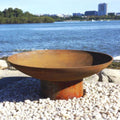 Rust-colored cast iron fire pit on pebbles by a waterfront with trees and buildings in the background.