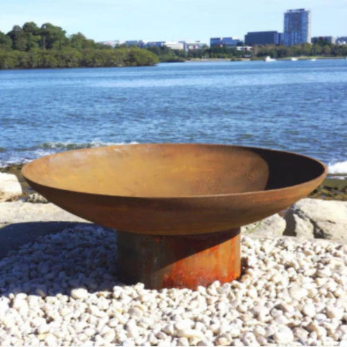 Rust-colored cast iron fire pit on pebbles by a waterfront with trees and buildings in the background.