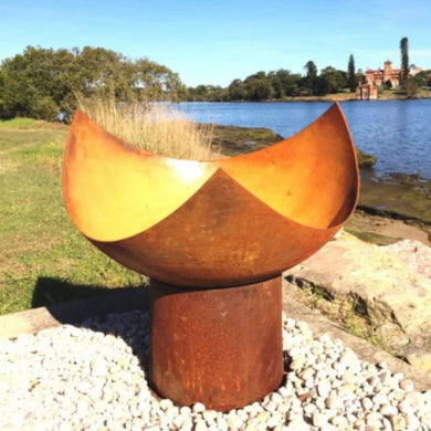 Cast Iron fire pit on pebbles near a body of water with trees and buildings in the background
