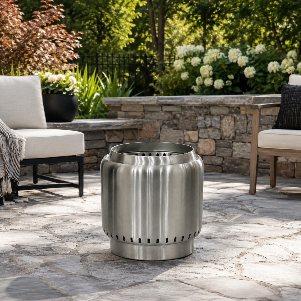 A smokeless stainless steel outdoor fire pit with a fluted cylindrical design sits on a stone patio in a backyard setting with patio furniture and greenery in the background.