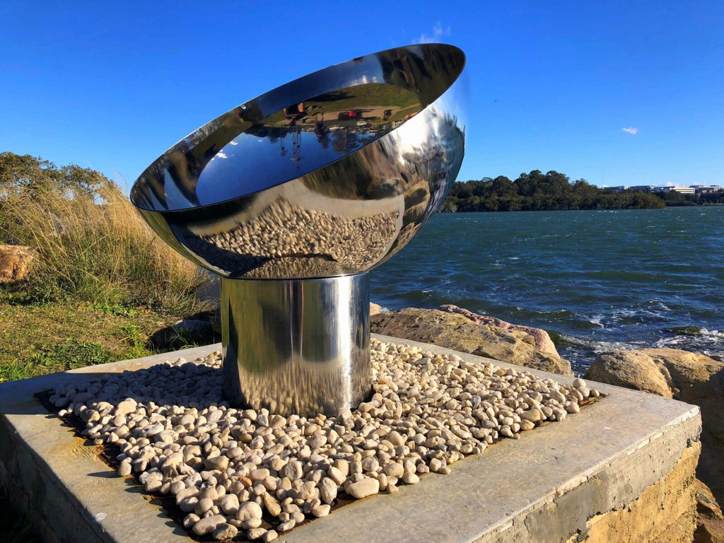 The Goblet Fire Pit – Stainless Steel on a pebble base by a waterfront with blue sky and water in the background