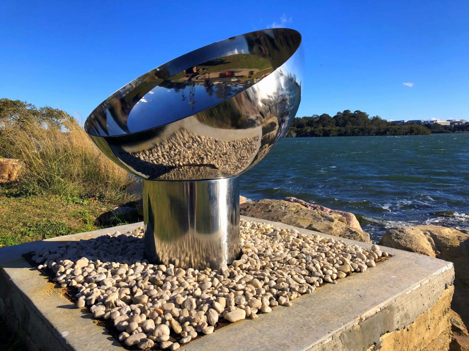 The Goblet Fire Pit – Stainless Steel on a pebble base by a waterfront with blue sky and water in the background