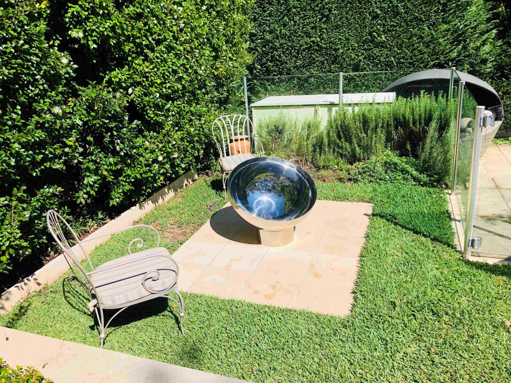 Garden scene with a large stainless steel fire pit on a stone platform, surrounded by greenery.
