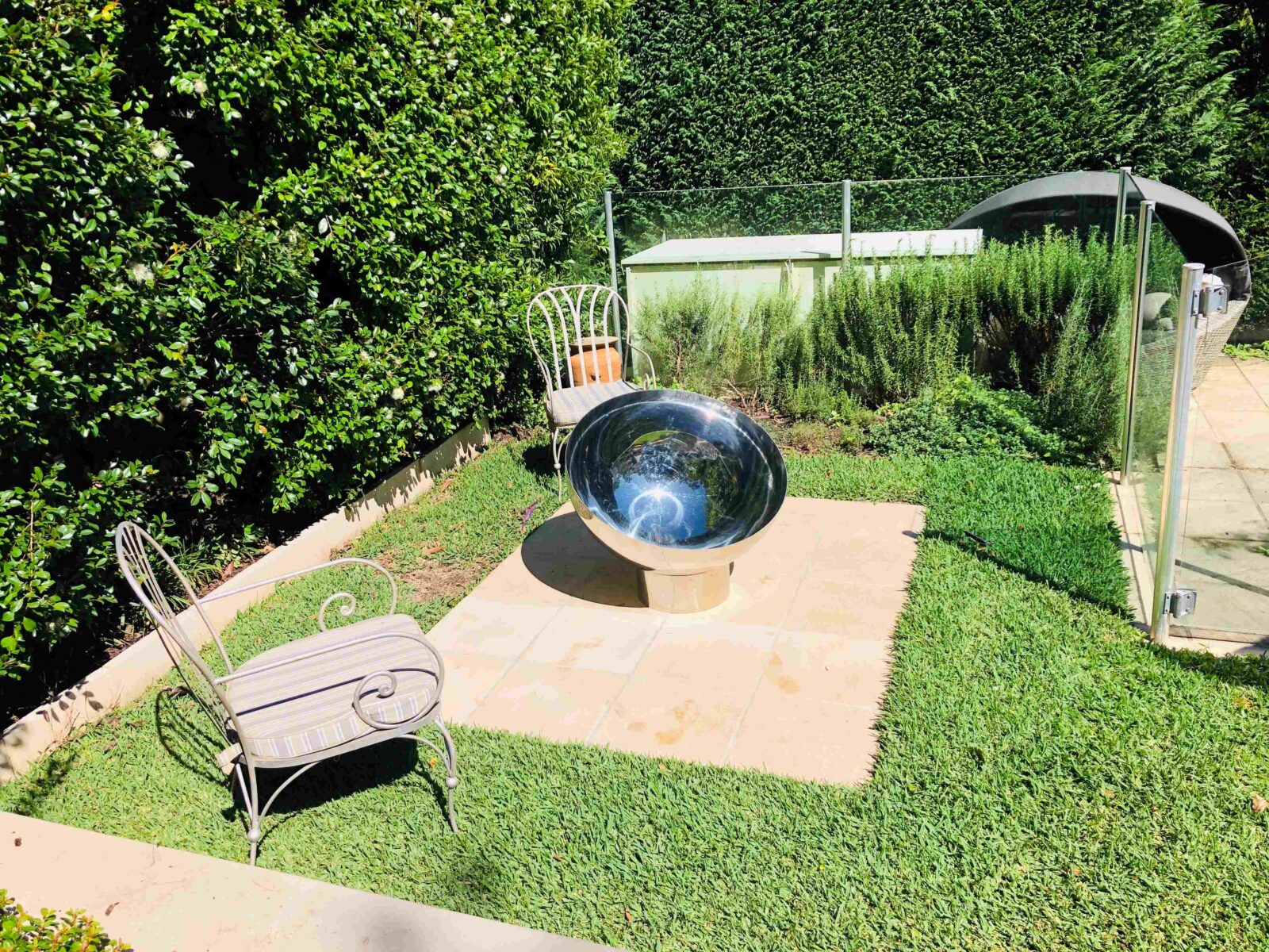 Garden scene with a large stainless steel fire pit on a stone platform, surrounded by greenery.