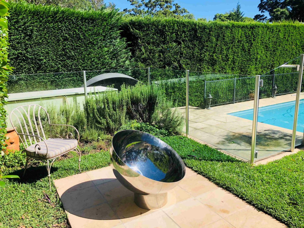 The Goblet Fire Pit – Stainless Steel on a patio with a pool and garden in the background
