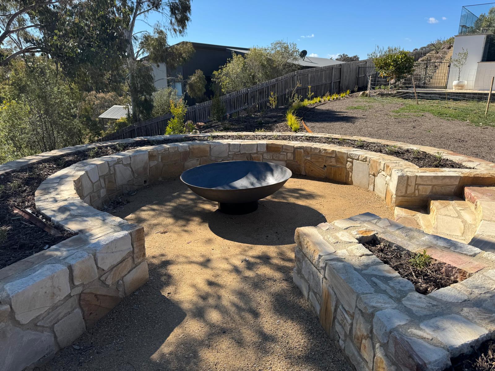 Outdoor stone fire pit seating area with a round cast iron fire pit centered inside curved stone walls on a sandy ground, with trees, fencing, and a house visible in the background.