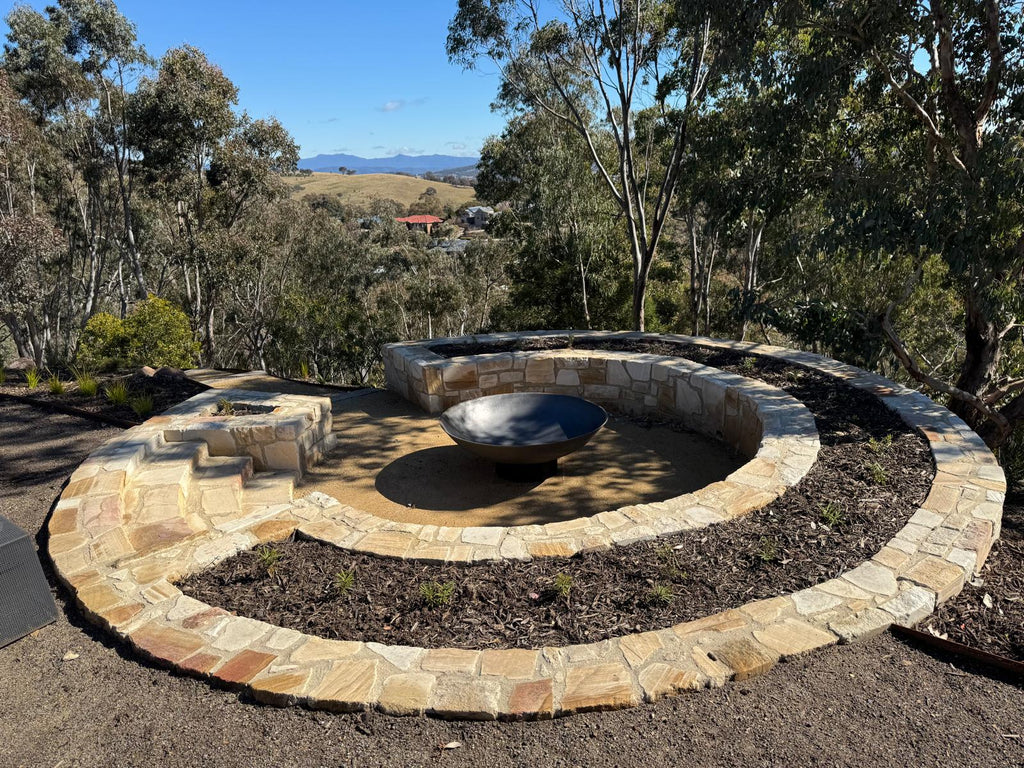 Outdoor stone fire pit seating arrangement with a circular stone border, built-in bench seating, and a cast iron fire pit centerpiece in a landscaped garden setting with trees and hills in the background.