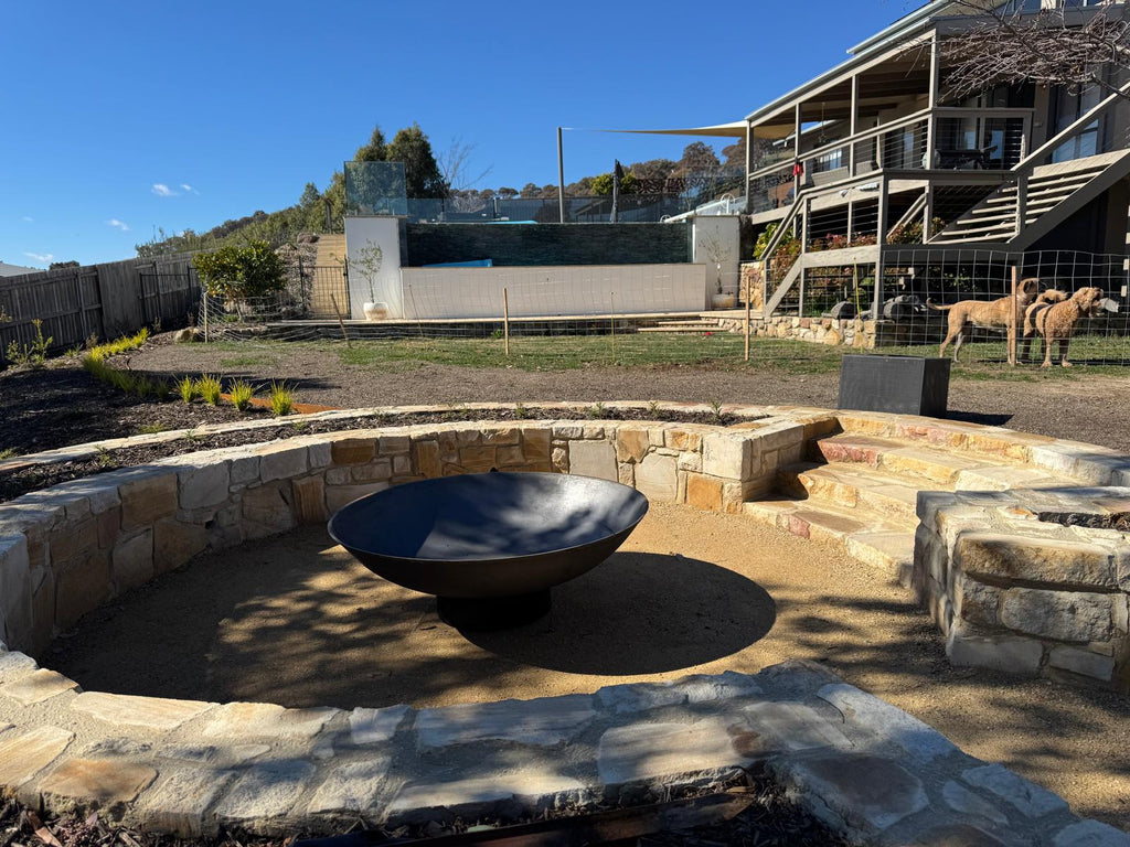 Outdoor stone fire pit area with a cast iron fire pit centerpiece, set in a backyard with a house, fence, and sheep visible in the background.