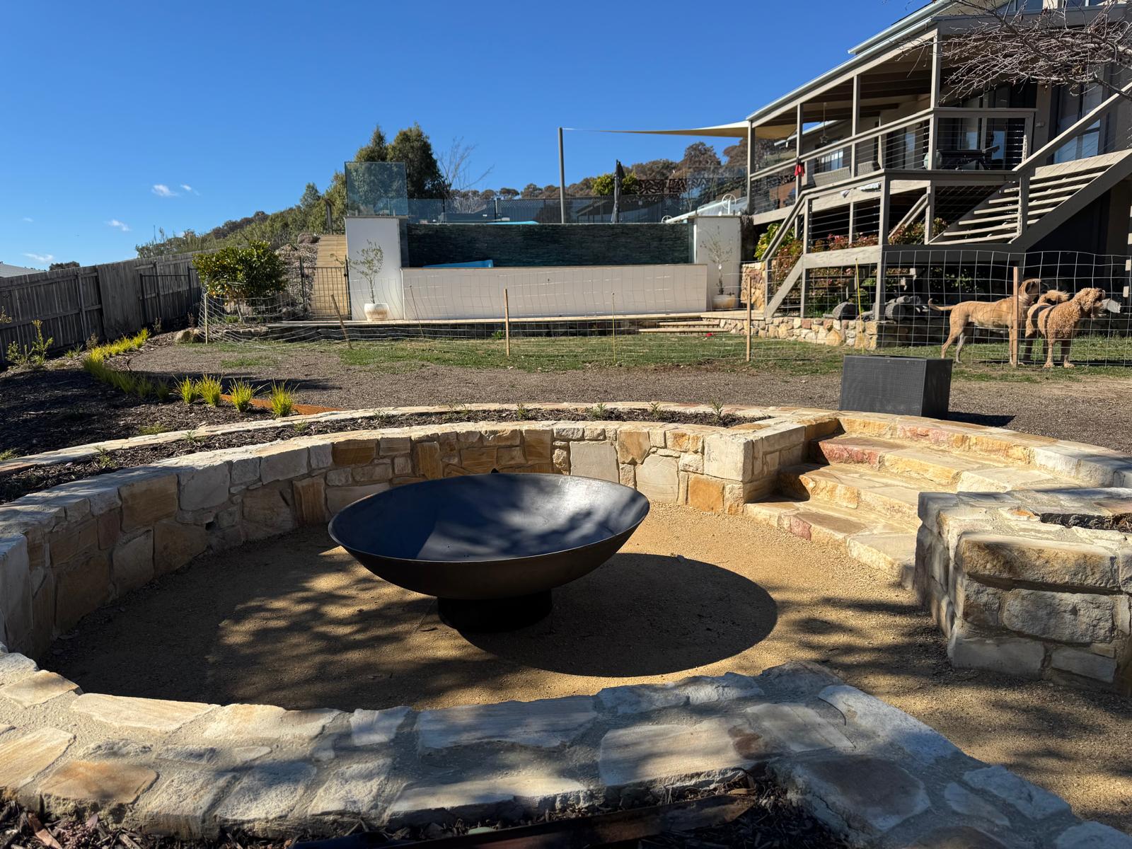 Outdoor stone fire pit area with a cast iron fire pit centerpiece, set in a backyard with a house, fence, and sheep visible in the background.