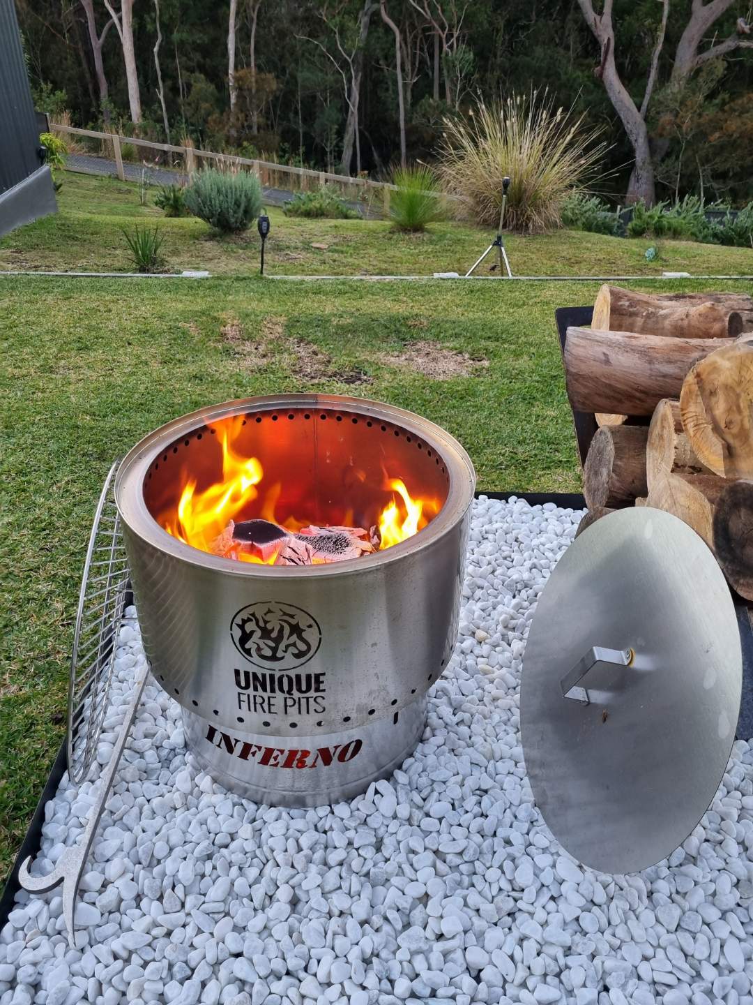 Fire pit with flames on a gravel surface in a garden setting, featuring 'Unique Fire Pits' branding.